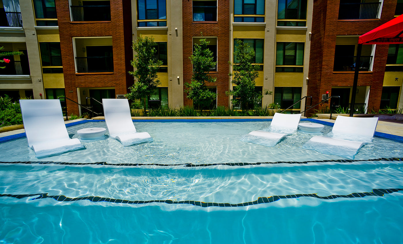 The Ledge Lounger Signature Chaise with Chairs and Side Table In-Pool 6-Piece Set sits partially submerged in a clear blue pool, with a modern apartment building and green trees visible in the background under bright sunlight.