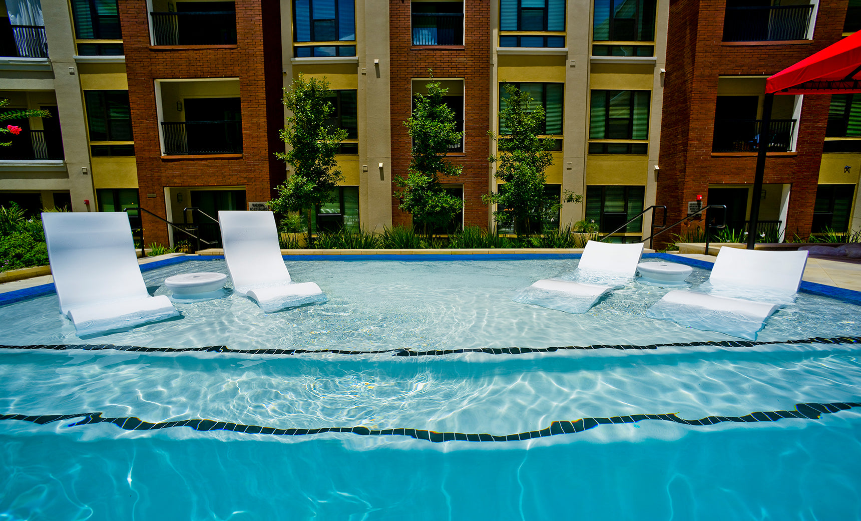 The Ledge Lounger Signature Chaise with Chairs and Side Table In-Pool 6-Piece Set sits partially submerged in a clear blue pool, with a modern apartment building and green trees visible in the background under bright sunlight.