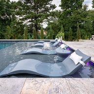A modern pool showcases the Ledge Lounger Signature Chaise and Side Table In-Pool 6-Piece Set, featuring gray chaises with white pillows and side tables. Poolside cushioned white loungers sit on a stone tile deck amid lush green trees for a resort vibe.