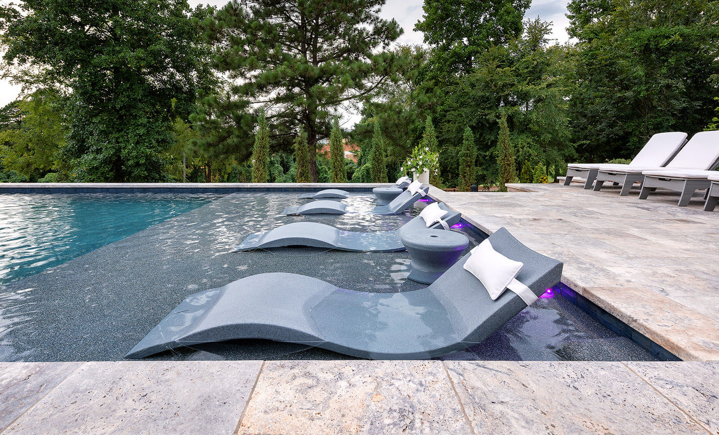A modern pool showcases the Ledge Lounger Signature Chaise and Side Table In-Pool 6-Piece Set, featuring gray chaises with white pillows and side tables. Poolside cushioned white loungers sit on a stone tile deck amid lush green trees for a resort vibe.