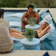 Three people enjoy the poolside: a man sits on the edge, another kneels in the water, and a woman relaxes on Ledge Lounger’s Signature Chaise and Side Table In-Pool 6-Piece Set, creating a laid-back summer vibe with drinks and potted plants nearby.