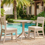The Ledge Lounger Mainstay Bistro 5-Piece Set, featuring cushioned chairs and a round table, is arranged poolside amid lush greenery and palm trees, creating an inviting spot to unwind with premium lounge furniture. Parts of a stone and stucco building are visible in the background.