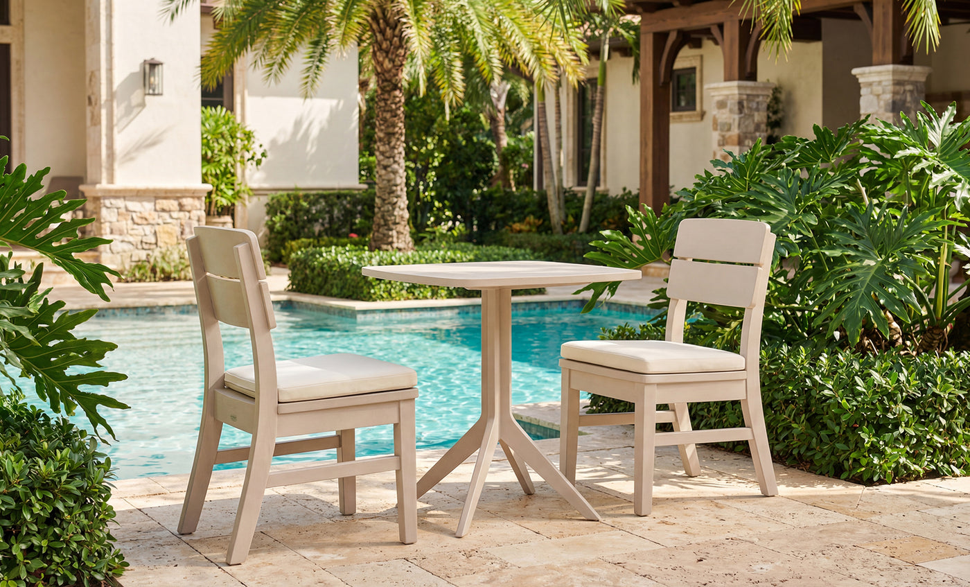 The Ledge Lounger Mainstay Bistro 5-Piece Set, featuring cushioned chairs and a round table, is arranged poolside amid lush greenery and palm trees, creating an inviting spot to unwind with premium lounge furniture. Parts of a stone and stucco building are visible in the background.
