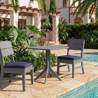 The Mainstay Bistro 5-Piece Set by Ledge Lounger features two gray chairs with navy cushions and a small table, elegantly arranged on a stone patio beside the pool. Surrounded by lush tropical plants, palm trees, and set against a Mediterranean-style building, this inviting ensemble provides the perfect setting to relax or entertain as your signature set for four.