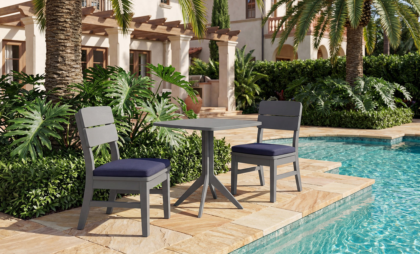 The Mainstay Bistro 5-Piece Set by Ledge Lounger features two gray chairs with navy cushions and a small table, elegantly arranged on a stone patio beside the pool. Surrounded by lush tropical plants, palm trees, and set against a Mediterranean-style building, this inviting ensemble provides the perfect setting to relax or entertain as your signature set for four.