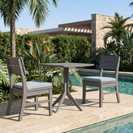 The Mainstay Bistro 5-Piece Set is arranged on stone tiles next to a clear swimming pool, surrounded by lush tropical plants, towering palm trees, and modern buildings in the background. Sleek Ledge Lounger poolside furniture and inviting in-pool chaises further enhance the scene, creating an ideal mix of relaxation and contemporary style.