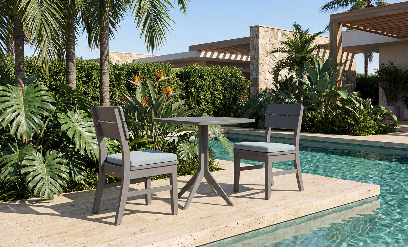 The Mainstay Bistro 5-Piece Set is arranged on stone tiles next to a clear swimming pool, surrounded by lush tropical plants, towering palm trees, and modern buildings in the background. Sleek Ledge Lounger poolside furniture and inviting in-pool chaises further enhance the scene, creating an ideal mix of relaxation and contemporary style.
