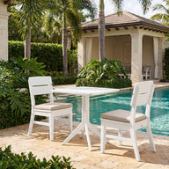 Two white dining chairs, each featuring a Mainstay Dining Side Chair Seat Cushion by Ledge Lounger for added comfort, are arranged with a square table on a stone patio next to a sparkling swimming pool. The setting is enhanced by lush green plants and palm trees, with a cabana and blue sky in the background.