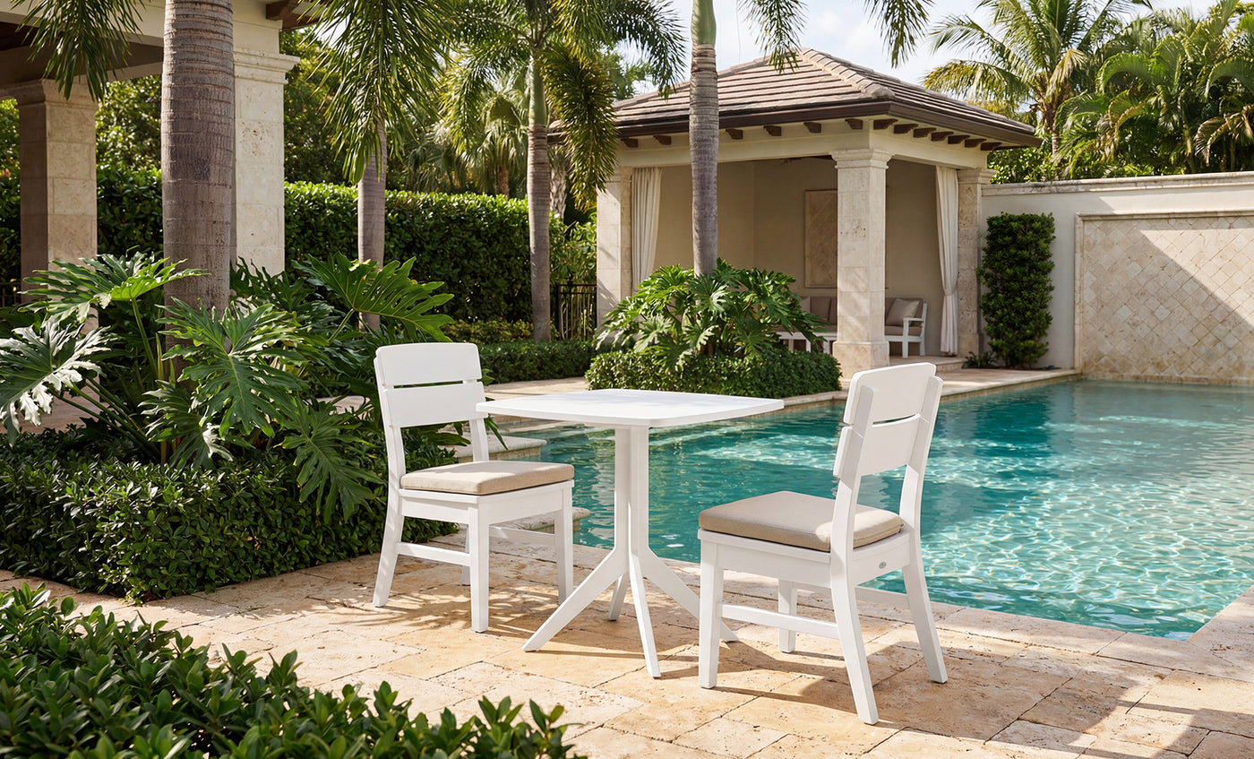 Two white dining chairs, each featuring a Mainstay Dining Side Chair Seat Cushion by Ledge Lounger for added comfort, are arranged with a square table on a stone patio next to a sparkling swimming pool. The setting is enhanced by lush green plants and palm trees, with a cabana and blue sky in the background.