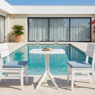 Two white chairs and a small table with wine and snacks are set beside a clear blue pool, surrounded by a modern house, blooming flowers, and potted plants on a sunny day—perfectly complemented by the Ledge Lounger Mainstay Bistro 5-Piece Set for ultimate relaxation.