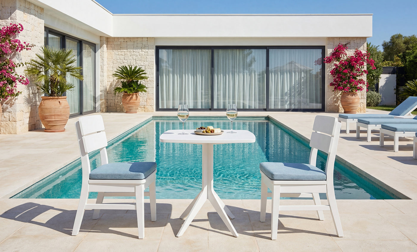 Two white chairs and a small table with wine and snacks are set beside a clear blue pool, surrounded by a modern house, blooming flowers, and potted plants on a sunny day—perfectly complemented by the Ledge Lounger Mainstay Bistro 5-Piece Set for ultimate relaxation.