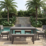 Transform your outdoor space with the Mainstay Dining 9-Piece Patio Set from Ledge Lounger. This elegant wooden set, featuring four chairs and a bench, is showcased poolside amid palm trees, lush greenery, and stylish stone paving under a partly cloudy sky. Perfect for al fresco gatherings or relaxing family meals, it blends durability and sophistication to elevate any patio setting.