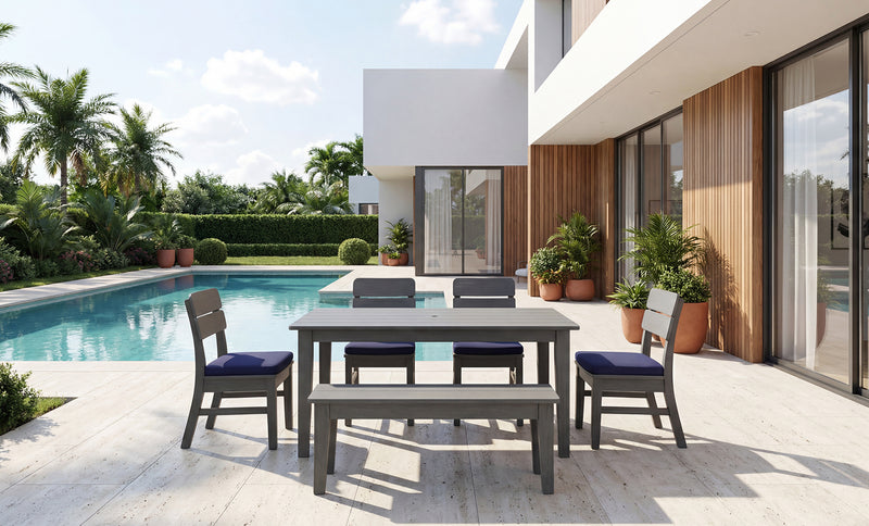 A 5-piece outdoor dining set with a rectangular gray table, matching bench, and four chairs with dark blue cushions, arranged on a light stone patio next to a swimming pool and modern house.