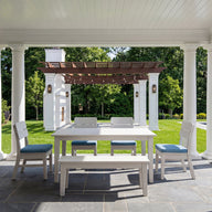 Experience outdoor elegance with the Mainstay Dining 9-Piece Patio Set by Ledge Lounger—a stunning ensemble featuring classic white columns, a spacious white dining table, and blue-cushioned chairs and bench. This inviting area overlooks a lush garden with a pergola and sparkling swimming pool, framed by green grass and trees on a sunny day. Ideal for those seeking inspiration or the perfect product description for your dream patio setup.