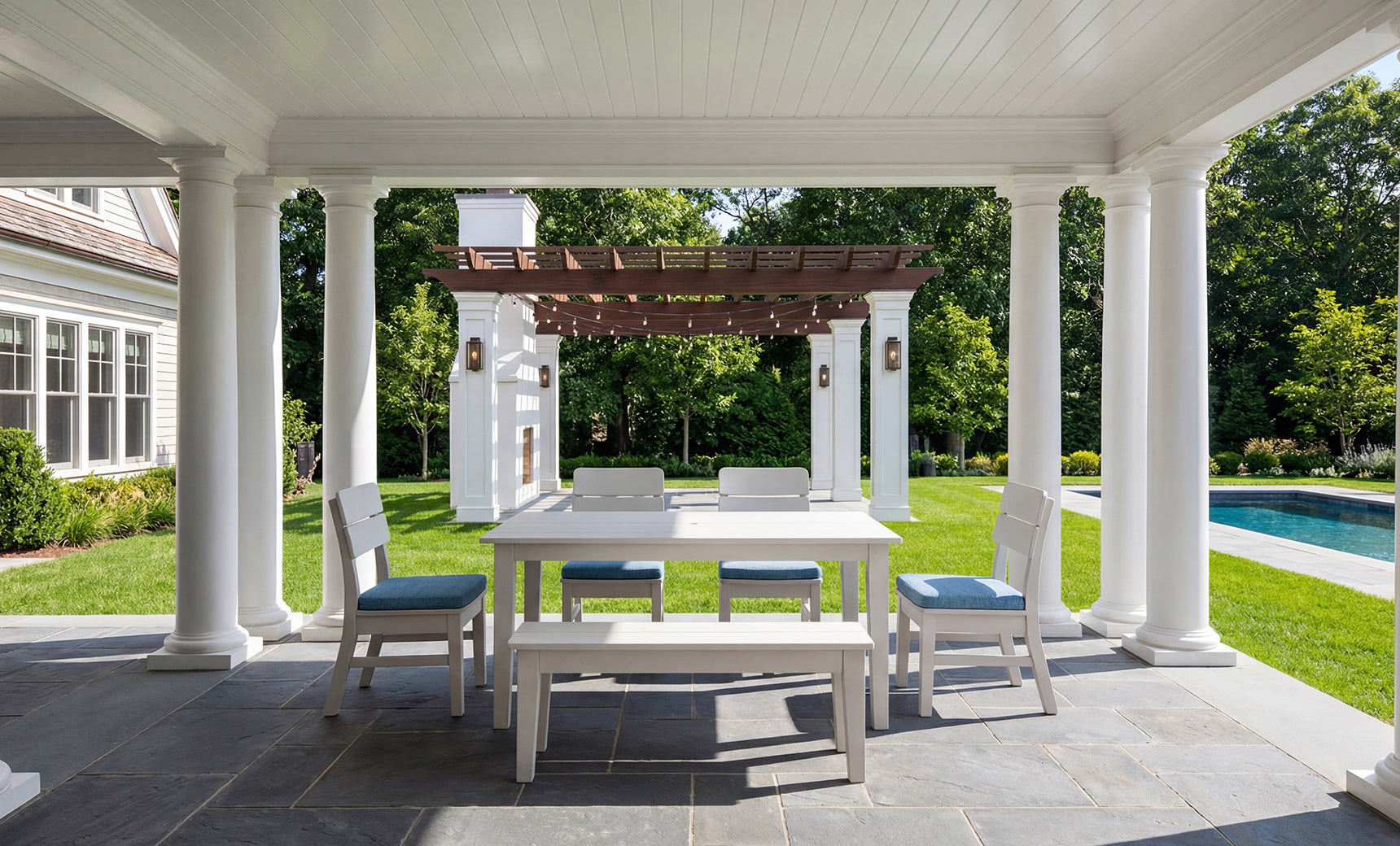 Experience outdoor elegance with the Mainstay Dining 9-Piece Patio Set by Ledge Lounger—a stunning ensemble featuring classic white columns, a spacious white dining table, and blue-cushioned chairs and bench. This inviting area overlooks a lush garden with a pergola and sparkling swimming pool, framed by green grass and trees on a sunny day. Ideal for those seeking inspiration or the perfect product description for your dream patio setup.