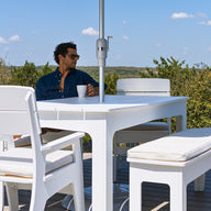 A person wearing sunglasses sits at a white outdoor table from the Mainstay Dining 9-Piece Patio Set by Ledge Lounger, surrounded by matching white chairs and a bench on a deck overlooking trees beneath a blue sky—an ideal setting for enjoying comfort and style outdoors.