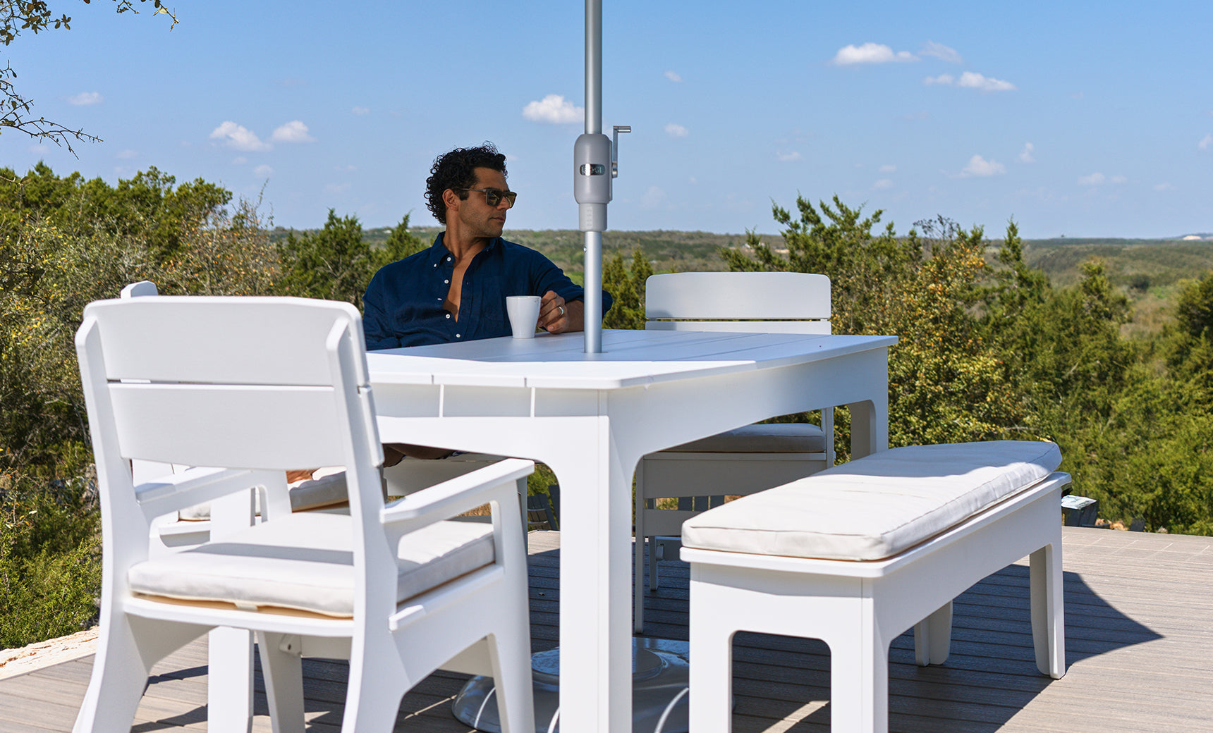 A person wearing sunglasses sits at a white outdoor table from the Mainstay Dining 9-Piece Patio Set by Ledge Lounger, surrounded by matching white chairs and a bench on a deck overlooking trees beneath a blue sky—an ideal setting for enjoying comfort and style outdoors.