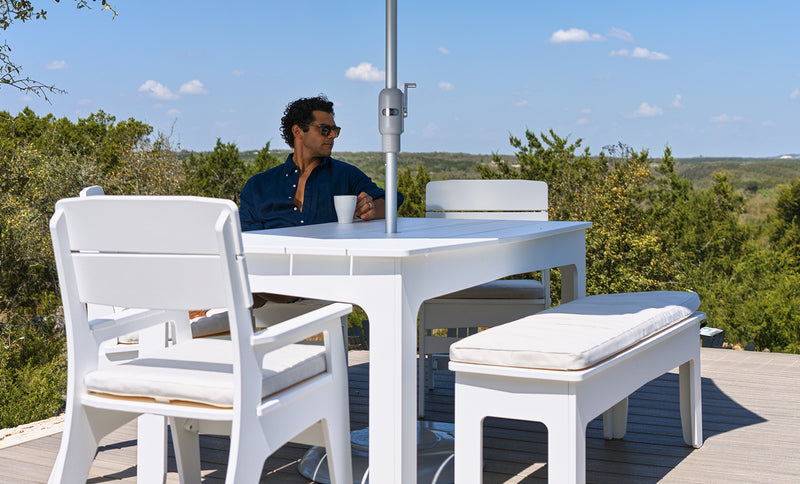 A person wearing sunglasses sits at a white outdoor table from the Mainstay Dining 9-Piece Patio Set by Ledge Lounger, surrounded by matching white chairs and a bench on a deck overlooking trees beneath a blue sky—an ideal setting for enjoying comfort and style outdoors.