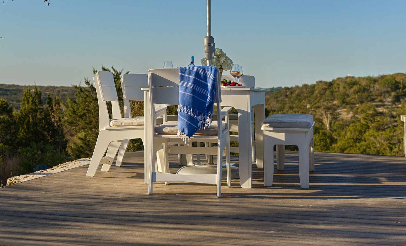 The Ledge Lounger Mainstay Dining 9-Piece Patio Set is showcased as a sample product, featuring a blue cloth draped over one chair and arranged on a wooden deck that overlooks scenic green trees and distant hills under a clear blue sky—making it an inviting addition to any outdoor space.