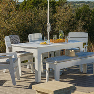 The Ledge Lounger Mainstay Dining 9-Piece Patio Set is arranged on a wooden deck, with chairs and a bench surrounding the table. Drinks and a hat sit atop the set, all framed by trees and rolling hills under a bright, sunny sky.