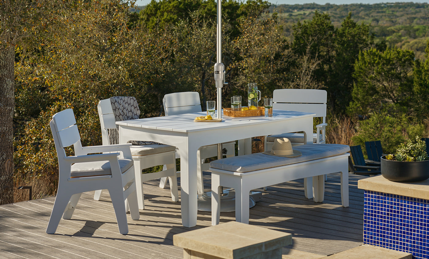 The Ledge Lounger Mainstay Dining 9-Piece Patio Set is arranged on a wooden deck, with chairs and a bench surrounding the table. Drinks and a hat sit atop the set, all framed by trees and rolling hills under a bright, sunny sky.
