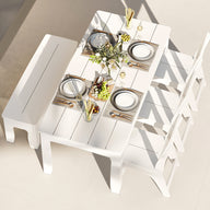 A bright, modern room features the Ledge Lounger Mainstay Dining 9-Piece Patio Set—a white dining table accompanied by three chairs and a bench, all set with plates, glasses, cutlery, a flower arrangement, grapes, and breadsticks—an inviting sample product that perfectly showcases contemporary dining elegance.