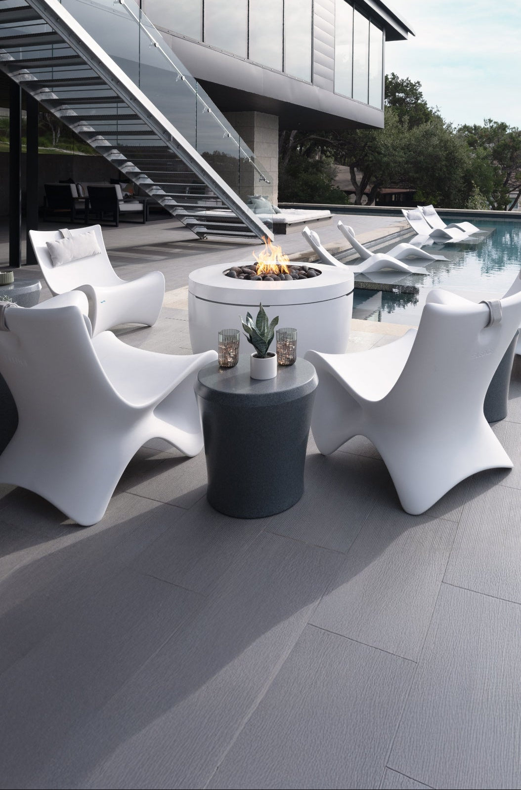 Modern outdoor setting with white chairs, a fire pit, and a pool in front of a modern building.