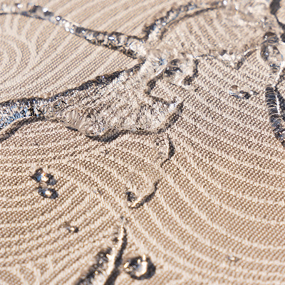 Close-up of water droplets beading on light beige, textured fabric with a wavy, circular pattern, showing the materials water-resistant properties.