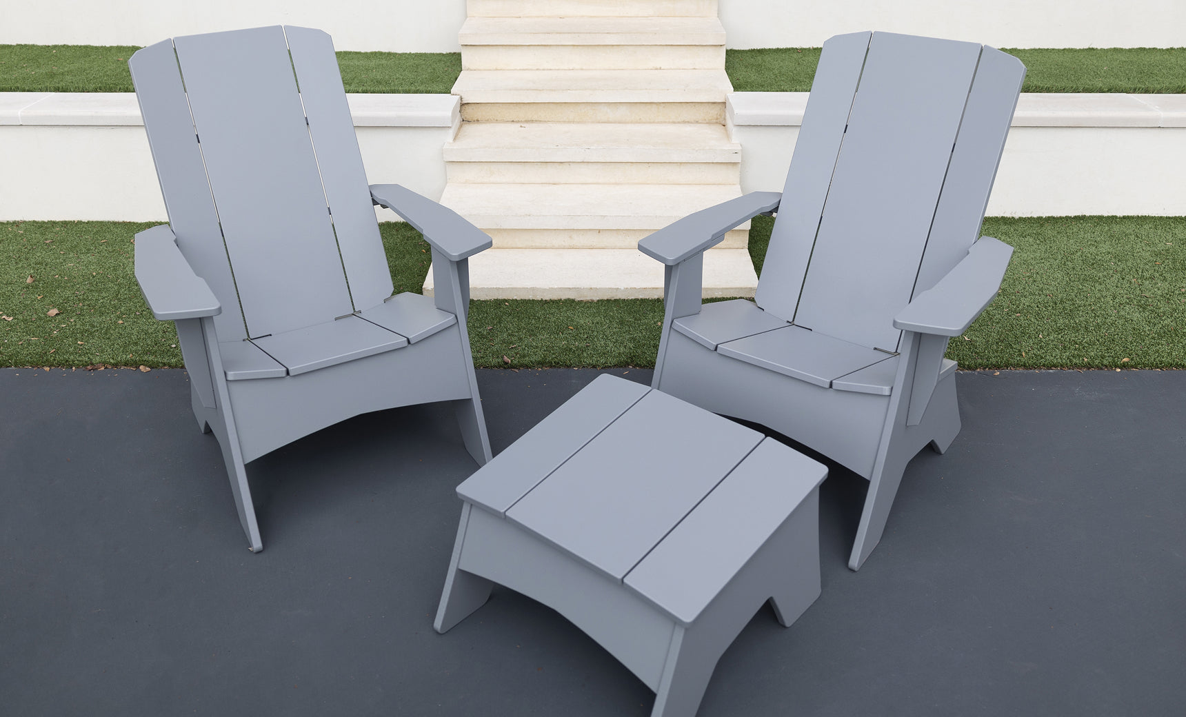 Two gray Ledge Lounger Mainstay Ultimate Adirondack chairs and a matching square table are set on a patio by white steps and artificial green grass.