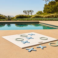 The Ledge Lounger Checkers & Tic Tac Toe set features oversized pieces on a poolside patio, surrounded by blue water, greenery, and a city view beneath a sunny sky.