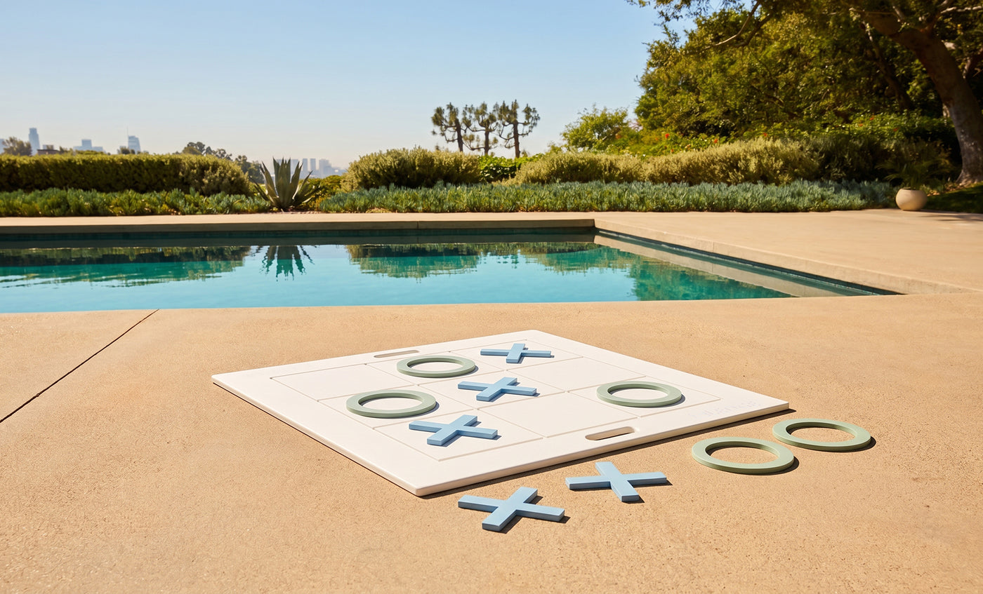 The Ledge Lounger Checkers & Tic Tac Toe set features oversized pieces on a poolside patio, surrounded by blue water, greenery, and a city view beneath a sunny sky.