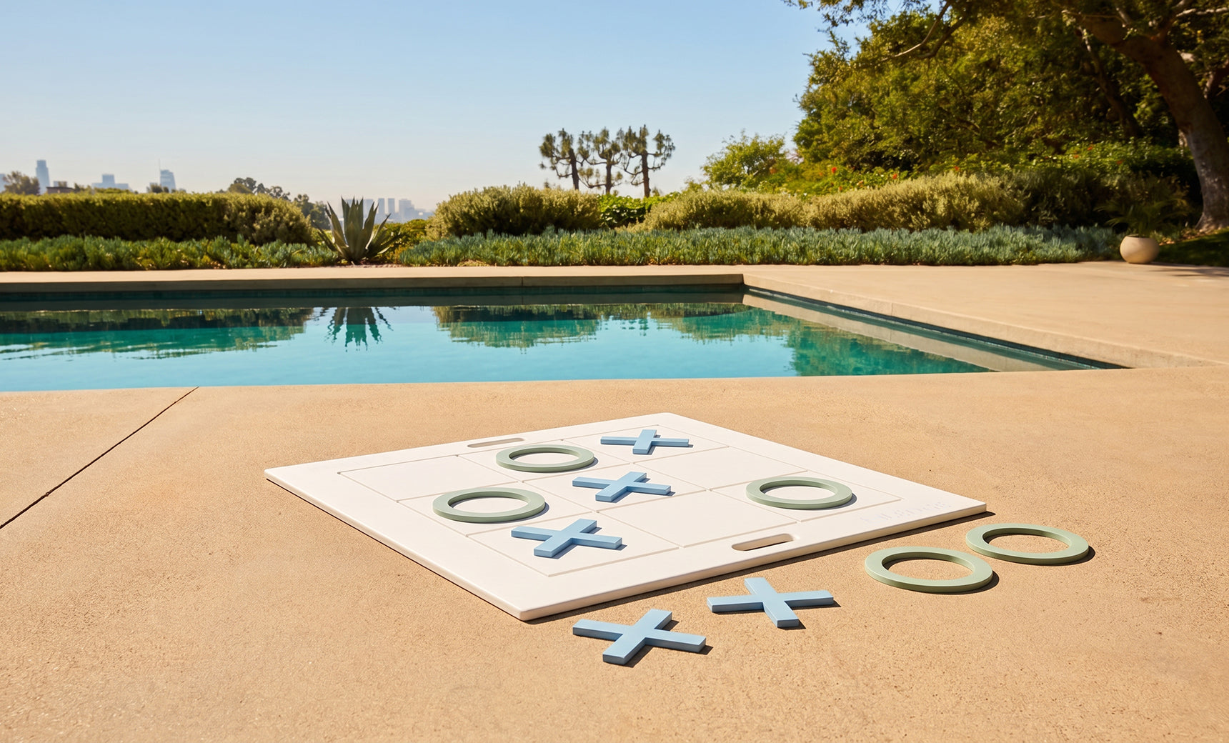 The Ledge Lounger Checkers & Tic Tac Toe set features oversized pieces on a poolside patio, surrounded by blue water, greenery, and a city view beneath a sunny sky.