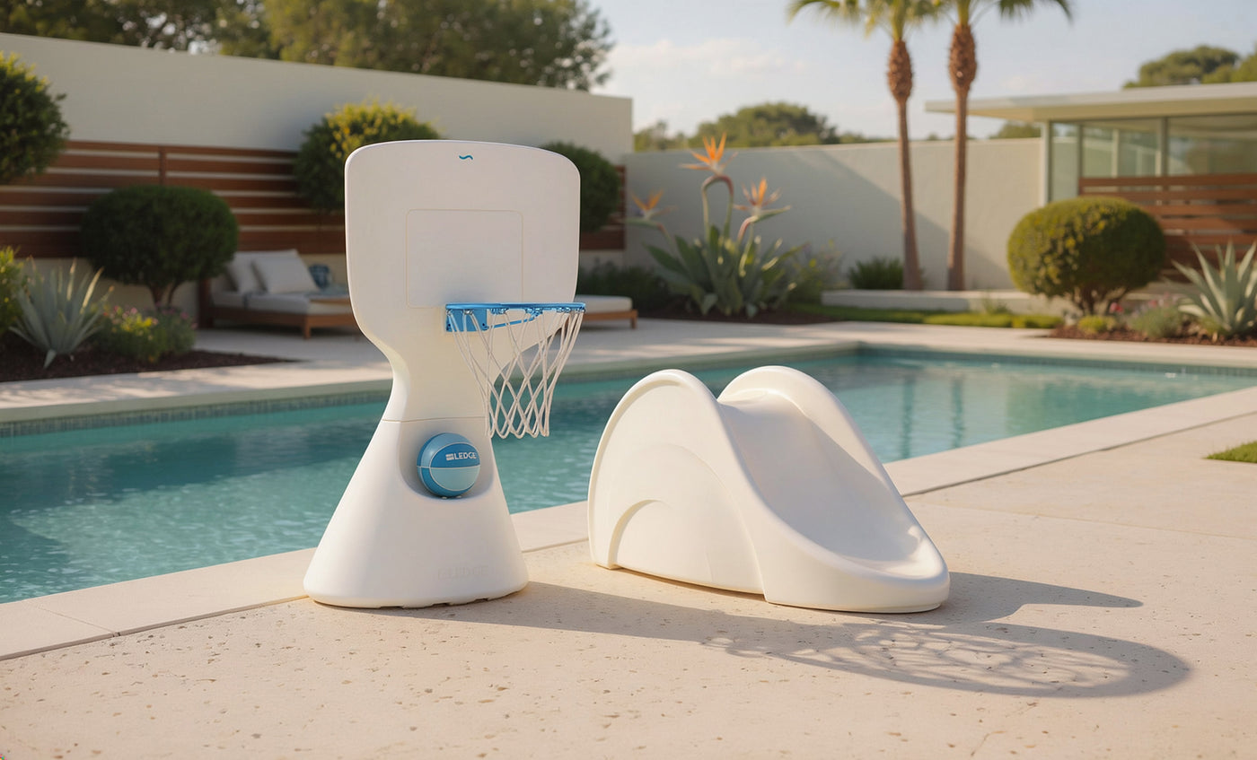 The Ledge Lounger Hoopstr and Signature Slide In-Pool Games Set are placed beside a modern outdoor pool with in-pool furniture, lounge chairs, and lush greenery, making for a fun and stylish setting on a sunny day.