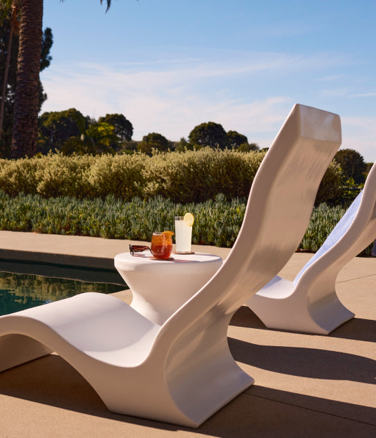 Two modern white lounge chairs by a pool with a small side table holding a cocktail, a clear drink with a lemon slice, and sunglasses. Lush greenery and trees are in the background under a partly cloudy sky.
