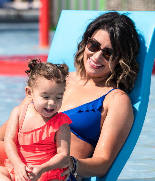A woman in sunglasses and a blue swimsuit smiles while holding a young girl in a bright orange swimsuit as they relax together on a poolside lounge chair.