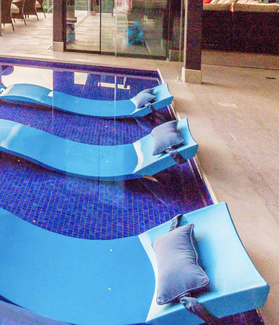 Three blue pool lounge chairs with gray pillows are partially submerged in a small, indoor pool with a tiled floor, near a seating area and large windows.