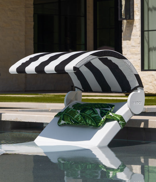 A white pool lounge chair sits in shallow water, topped with a black-and-white striped canopy and green leaf-print pillows, with a modern building and grass in the background.