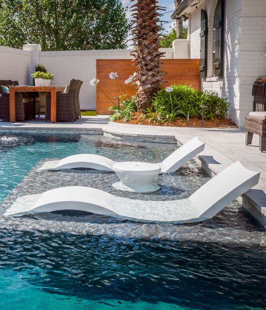 Two white lounge chairs and a small table sit on a shallow ledge in a modern backyard pool. A dining area, palm tree, and lush landscaping are visible in the background.