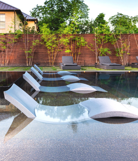 Five modern lounge chairs are partially submerged in a shallow pool, lined up facing a wooden privacy fence with green bamboo plants and poolside chairs in the background.