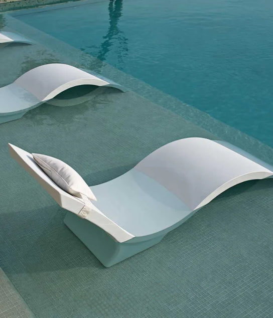 A modern white pool lounge chair with a pillow is partially submerged in shallow water, next to similar chairs, creating a relaxing poolside atmosphere.