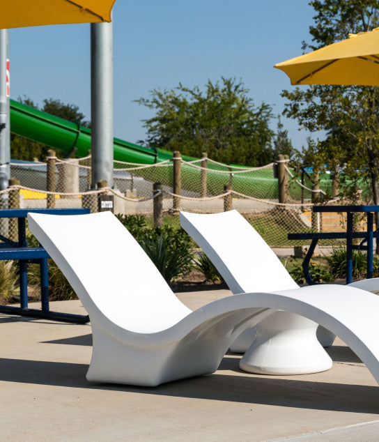 Two modern white lounge chairs sit by a poolside under yellow umbrellas, with a green water slide and trees visible in the background on a sunny day.