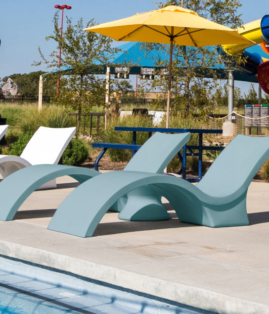 Curved lounge chairs sit poolside near a bright yellow umbrella, with water slides and greenery in the background at an outdoor recreational area.
