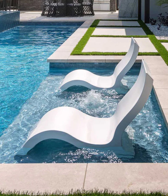 Two modern white lounge chairs sit partially submerged in the shallow end of a stylish swimming pool with clear water and geometric poolside tiles.
