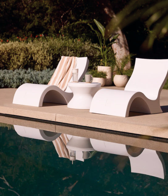 Two modern white lounge chairs and a small round table with cups sit by a pool, reflecting in the water. A towel hangs on one chair. Lush greenery is visible in the background.