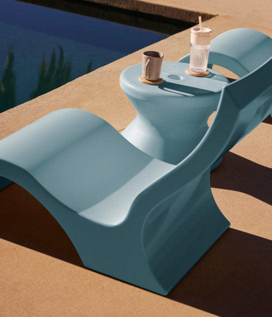 A modern blue poolside lounge chair and table set on tan concrete. The table holds a mug, a glass of water with a straw, and another mug. The pools edge and water are visible in the background.