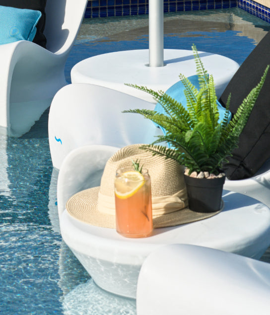 A straw hat, a glass of iced tea with lemon, and a potted fern rest on a white poolside table beside lounge chairs in a shallow pool.