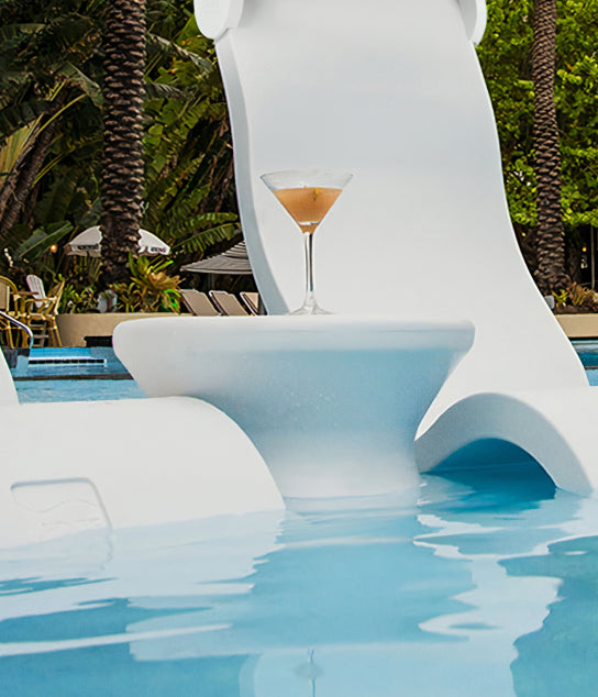 A martini glass sits on a white table in a pool, near a water slide, with palm trees and lounge chairs in the background.