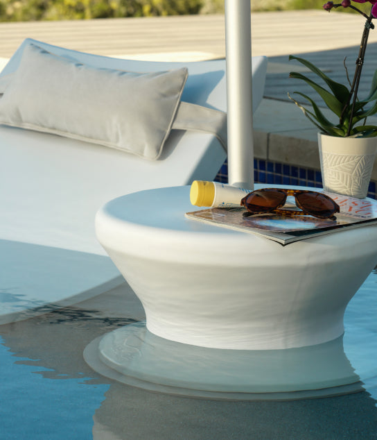 A lounge chair beside a pool with a pillow behind it. On a nearby white table are sunglasses, a tube of sunscreen, a magazine, and a potted plant. The table sits partially in the water.