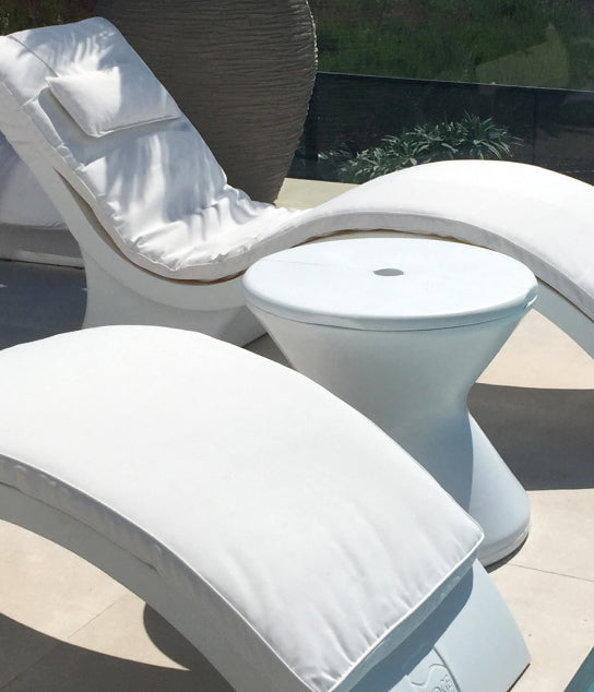 White curved outdoor lounge chairs with cushions and a small round white side table are arranged on a sunny patio. The background includes greenery and a glass barrier.