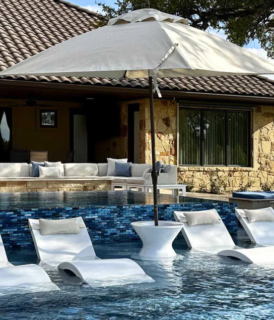 White lounge chairs and a large umbrella sit partially submerged in a swimming pool, with a stone house and a shaded outdoor seating area in the background. Blue mosaic tiles border the pool edge.